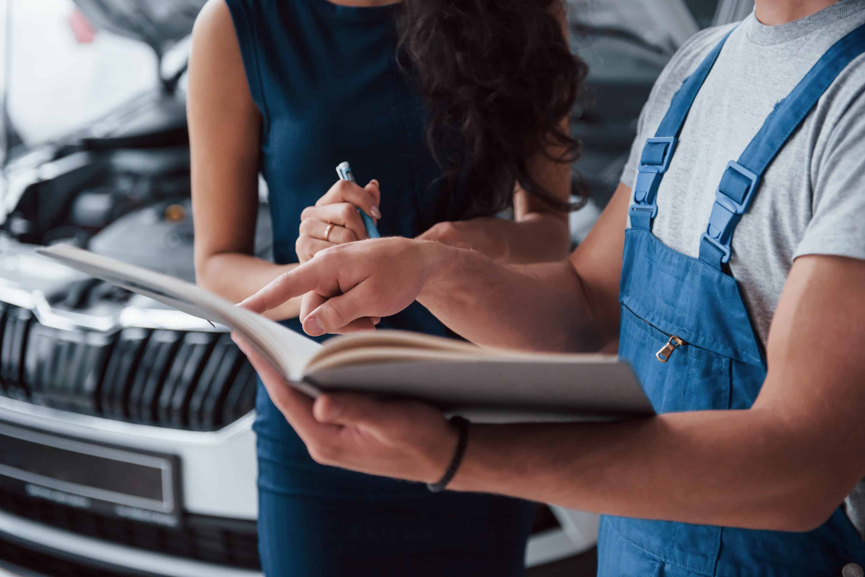 Ein Spezialist für Unfallreparatur beratet eine Kundin. Zu sehen ist ein Mann im Arbeitsanzug mit Stift und Notizbuch neben einer Frau in schwarzem Kleid mit einem dunklen Auto im Hintergrund.