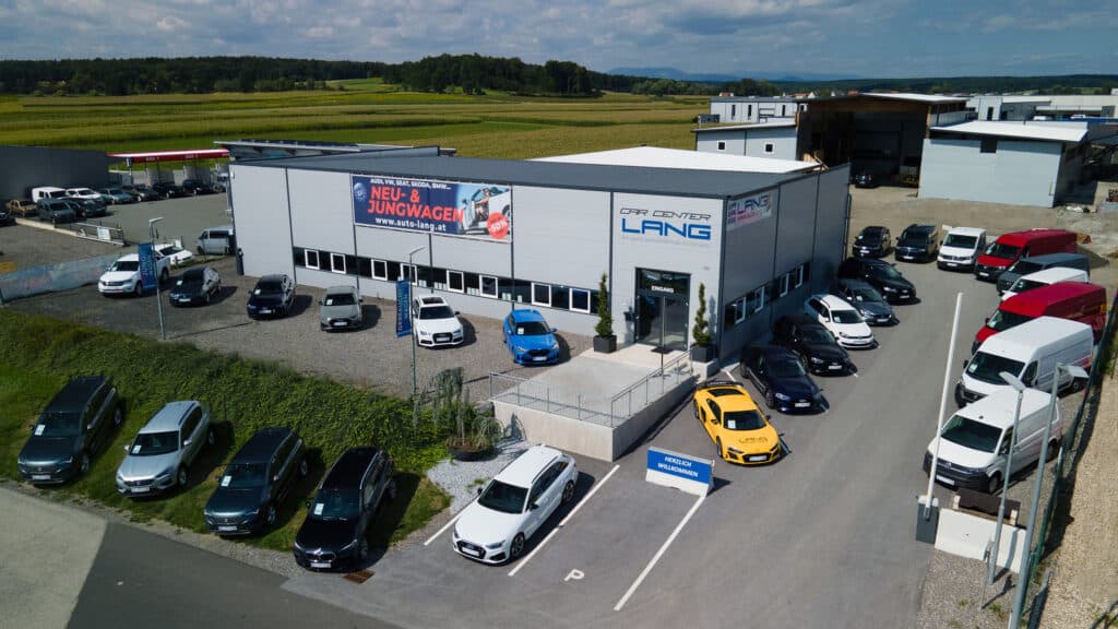 Ein Blick auf das Car Center Ilz von der Vogelperspektive aus, ausgehend von Richtung Autobahnabfahrt. Man sieht eine graue Halle mit LANG Logo, mehreren Parkplätzen und diversen Fahrzeugen, die vor der Halle parken.