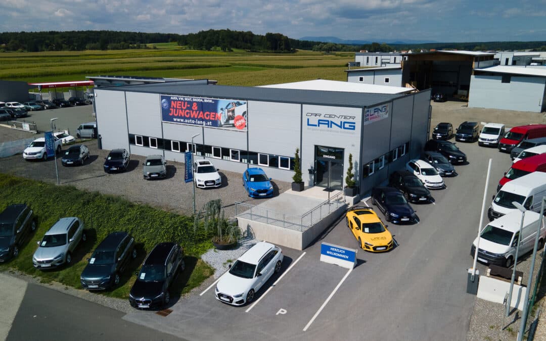 Ein Blick auf das Car Center Ilz von der Vogelperspektive aus, ausgehend von Richtung Autobahnabfahrt. Man sieht eine graue Halle mit LANG Logo, mehreren Parkplätzen und diversen Fahrzeugen, die vor der Halle parken.