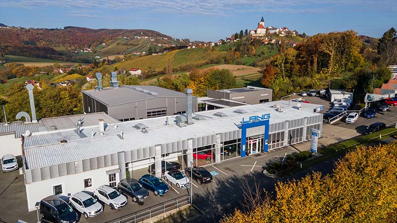 Eine Luftaufnahme zeigt das Autohaus Lang mit Verkaufshalle, dahinter das neue Karosserie- und Lackiercenter und hinter den Gebäuden sieht man den Ort Sankt Anna am Aigen.