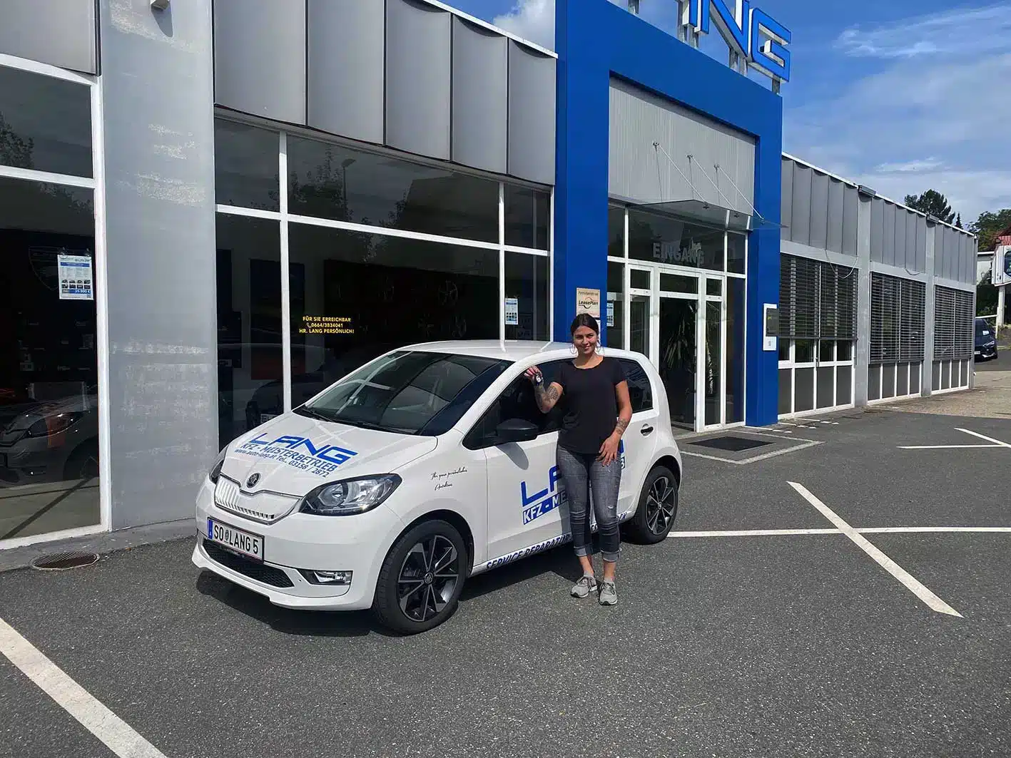 Eine Frau steht neben einem Fahrzeug, welches mit dem Autohaus Lang Logo foliert ist. Im Hintergrund sieht man das Autohaus Lang, welches eine graue Fassade hat und dessen Eingang ein blaues Tor mit einem großen LANG darüber ist. 